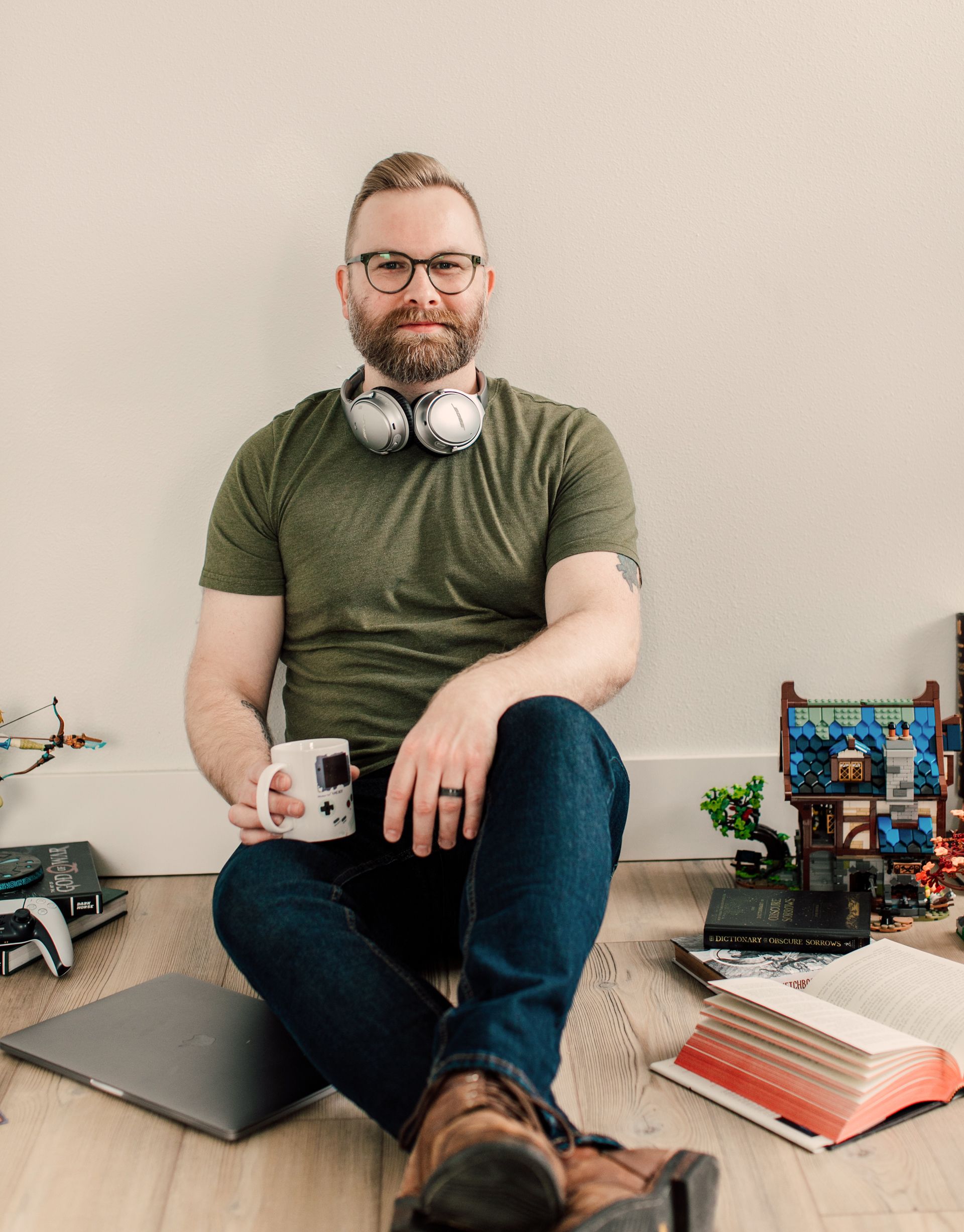 a portrait of Kyle Jorve, holding a coffee mug with a Game Boy graphic, sitting against a wall, with books and LEGO sets scattered around him